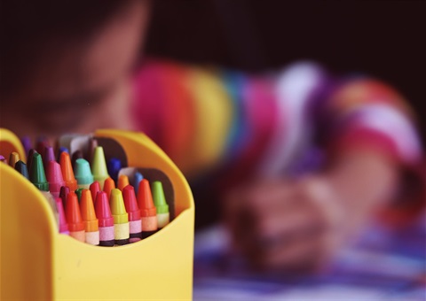 Crayons in a box in focus and a child drawing in the background